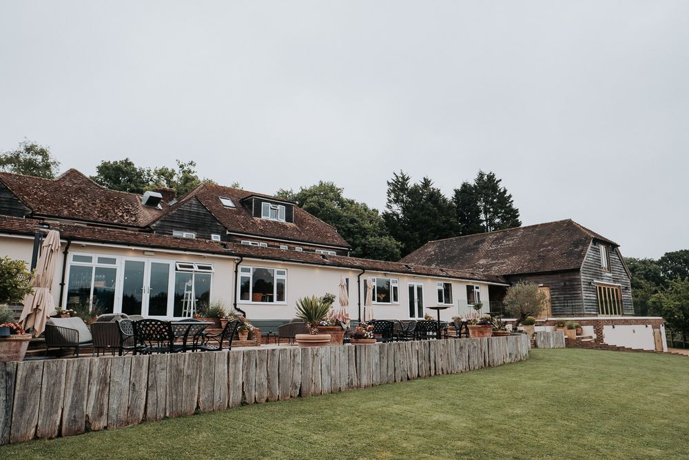 Charming English countryside barn venue with wooden fence, manicured lawn, and patio seating under an overcast sky.