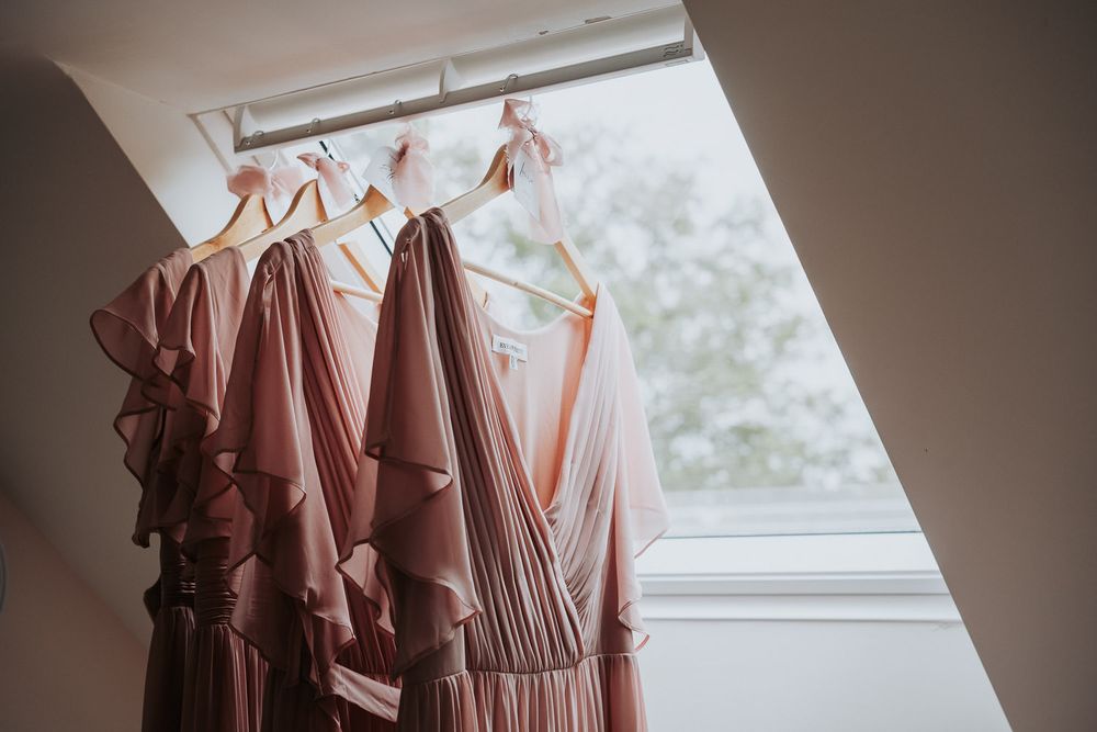 Pink bridesmaid dresses hanging on wooden hangers near a skylight window in soft natural light.