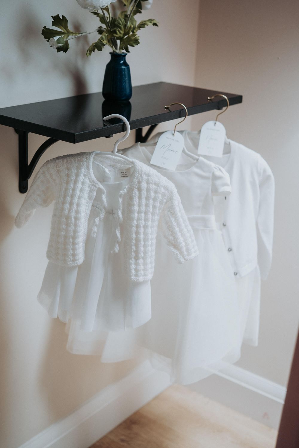 Two white baby dresses hanging on hangers beneath a black shelf with a blue vase and flowers.