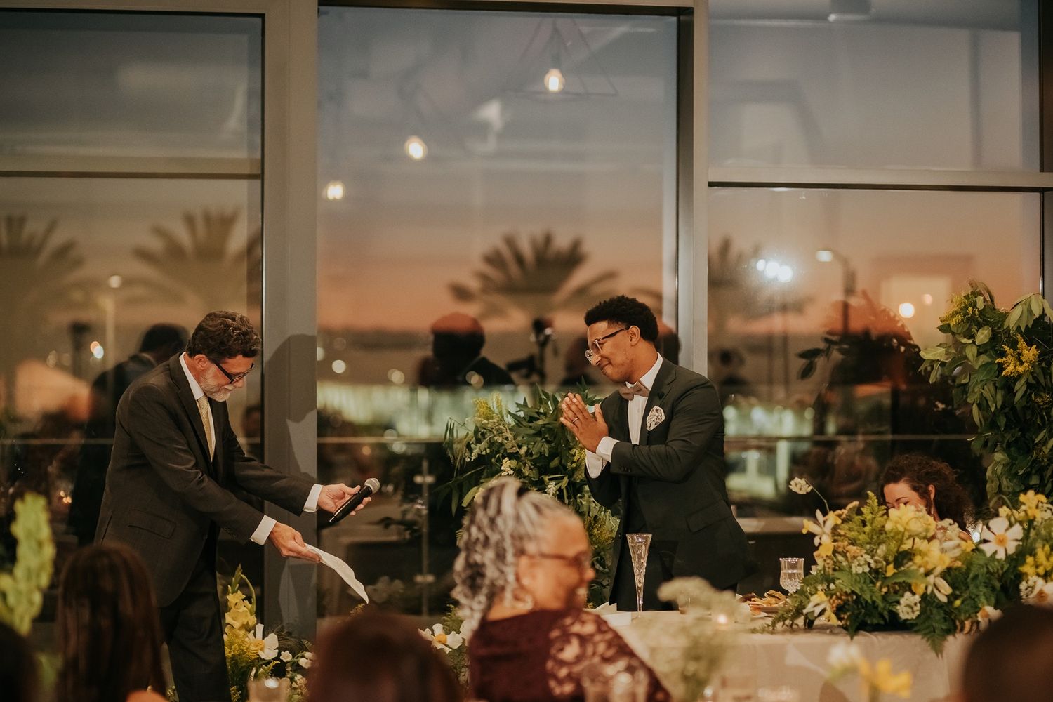 The groom and a guest share a joyful moment during their wedding reception with floral arrangements and a sunset view at The Lane.