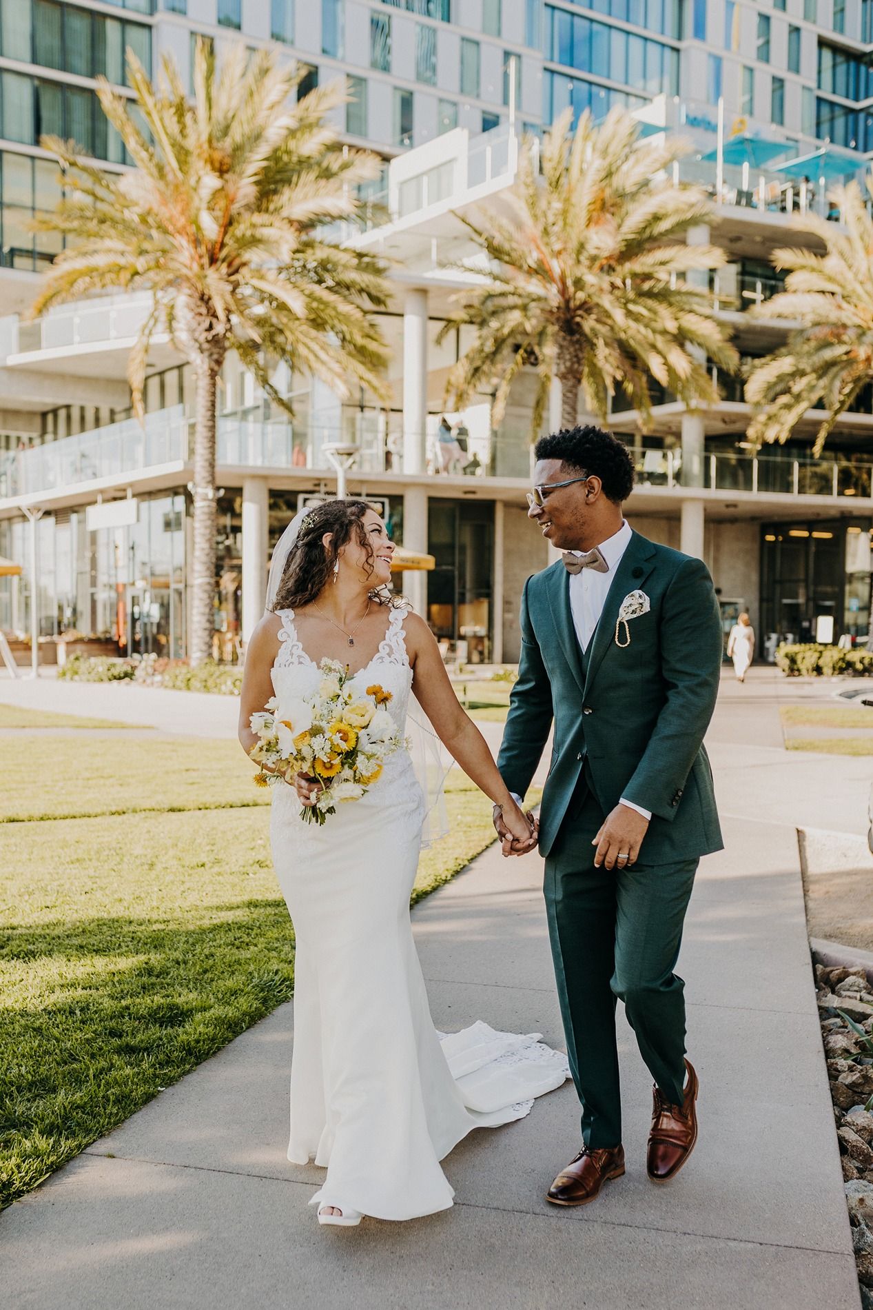 A bride in a white gown and groom in a green suit hold hands walking past palm trees on their wedding day at The Lane.