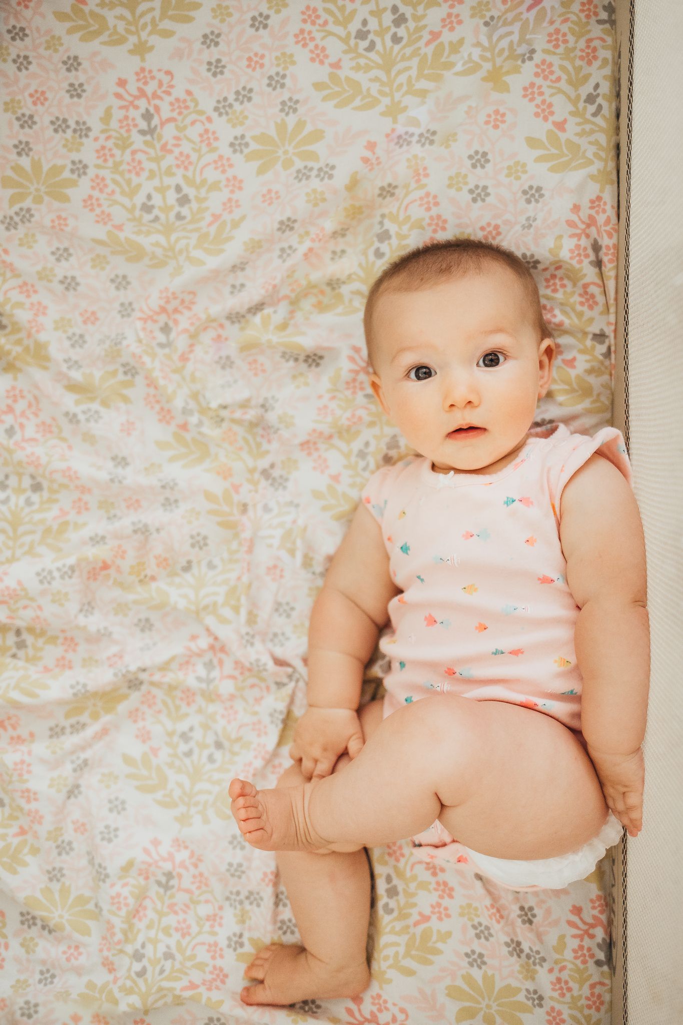 Adorable baby girl in pink onesie lying on floral patterned blanket, looking up with curious eyes.