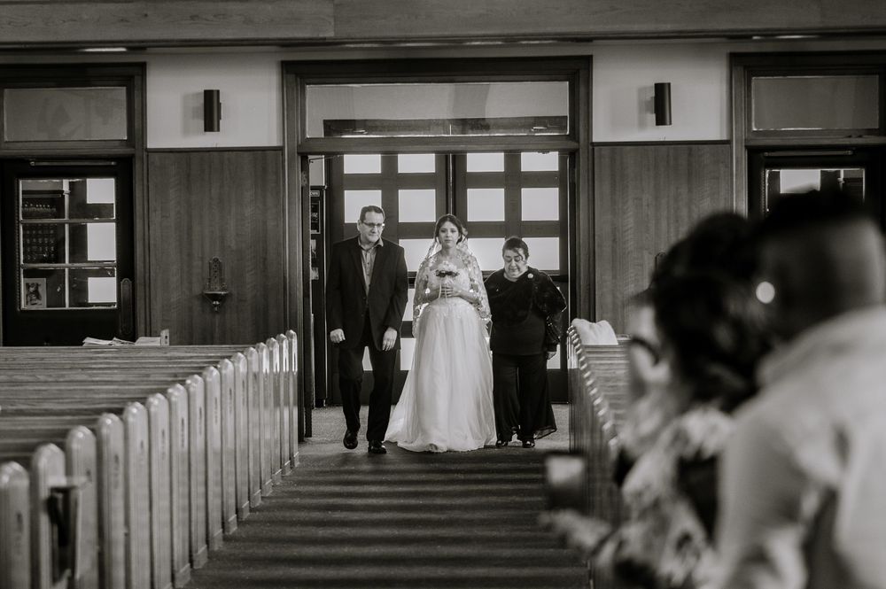 Black and white wedding photo of bride walking down church aisle escorted by two people, with pews and guests visible.