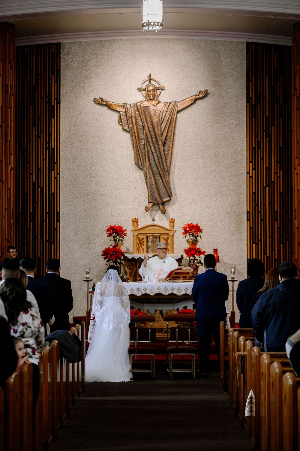A bride and groom stand at the altar during a Catholic wedding ceremony beneath a golden Christ statue.