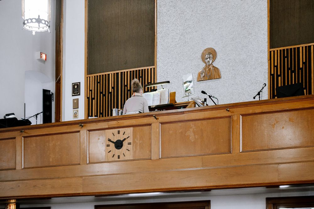 Person seated at a wooden balcony pulpit with clock, microphone, and religious portrait in a church interior.