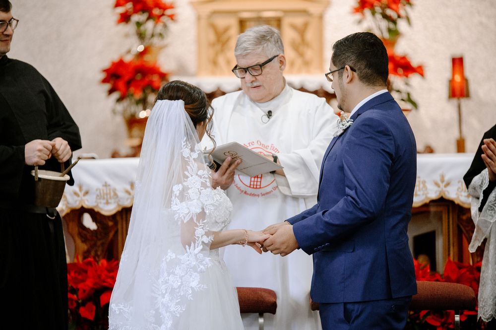 Bride and groom exchange vows at a church wedding ceremony with a officiant holding a book near a festive altar.