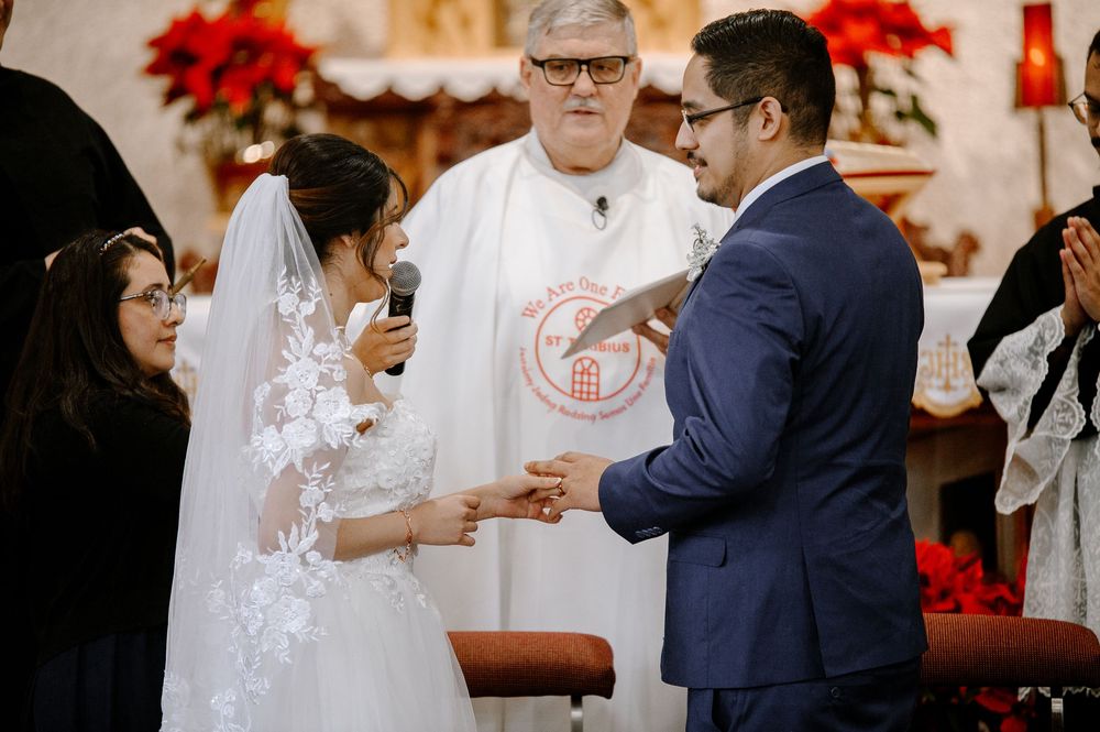 Bride and groom exchange vows at church wedding ceremony with officiant in white robe and holiday floral decorations.