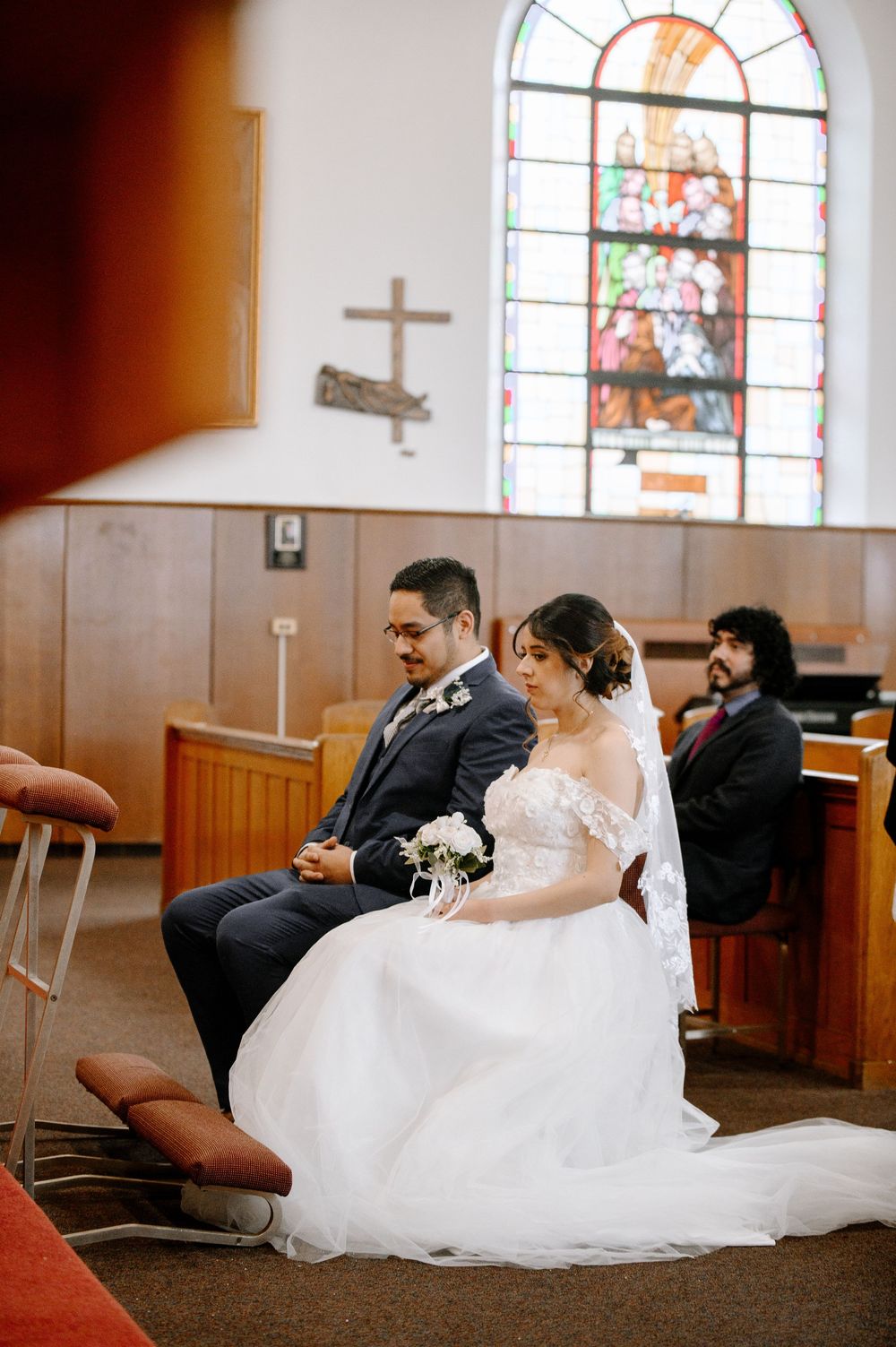 Bride and groom sit together in church pews during wedding ceremony, with stained glass window and cross in background.