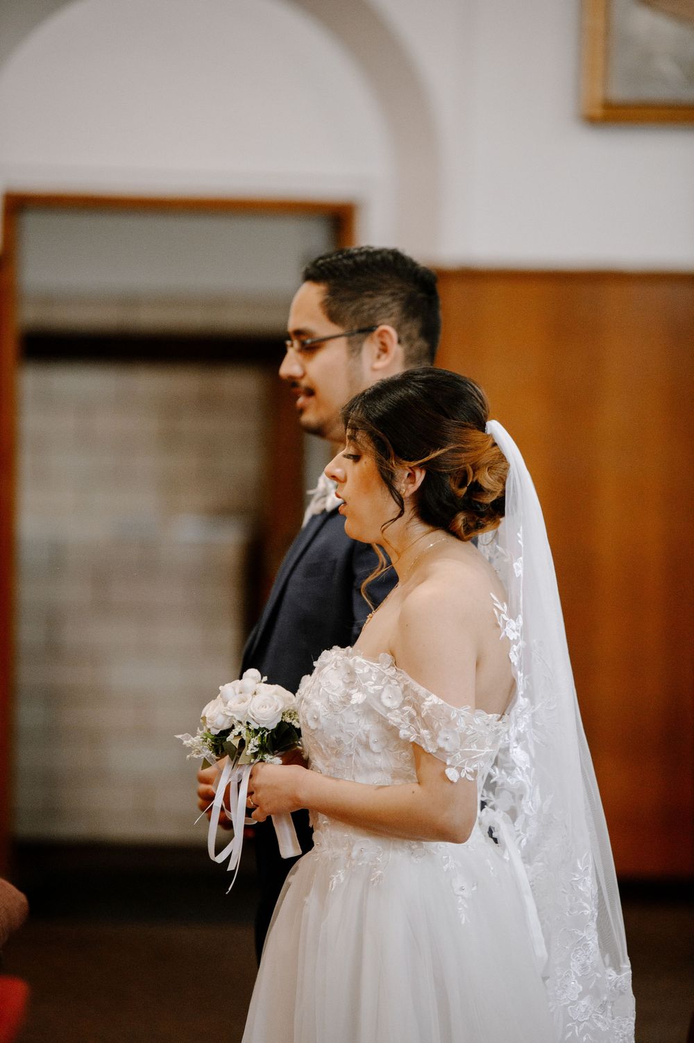 Bride in lace off-shoulder gown holding white bouquet stands beside groom in dark suit during church wedding ceremony.
