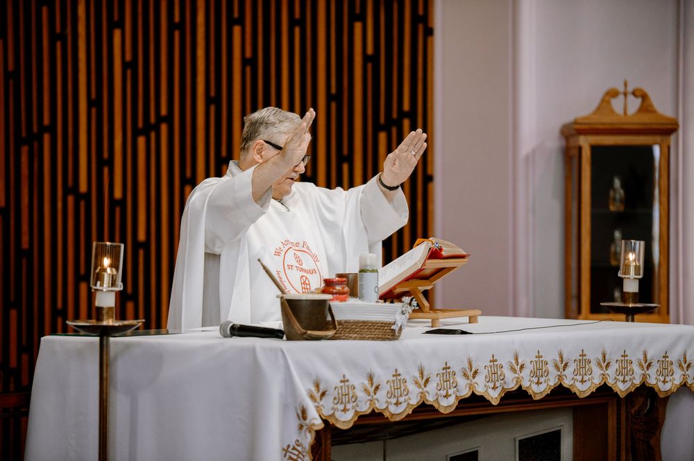 Catholic priest in white vestments raises hands during Mass at an altar with candles and open Bible.