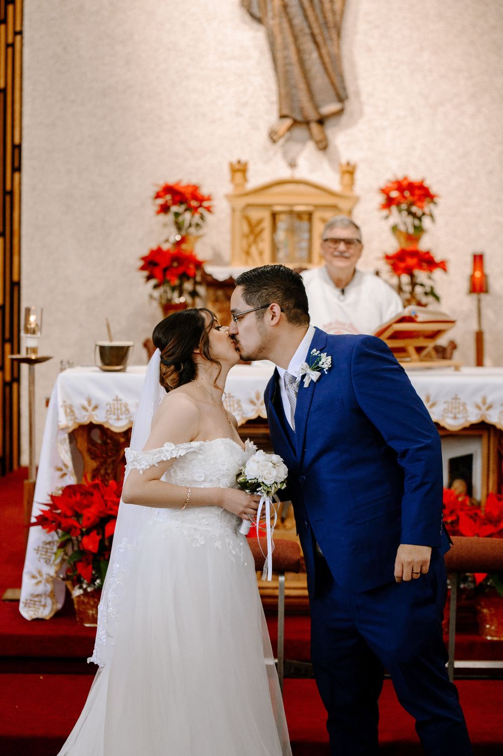 Bride and groom share first kiss at church altar adorned with red poinsettias, officiant smiling behind them.