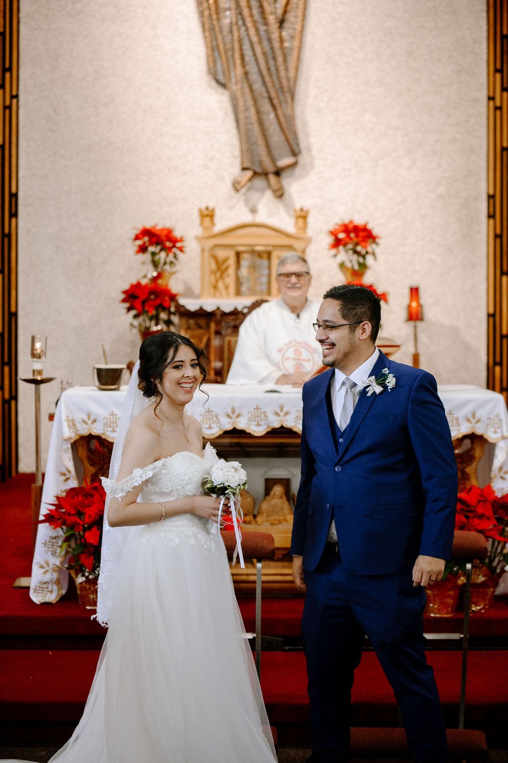 Bride in white gown and groom in blue suit smile at altar adorned with red poinsettias during church wedding ceremony.