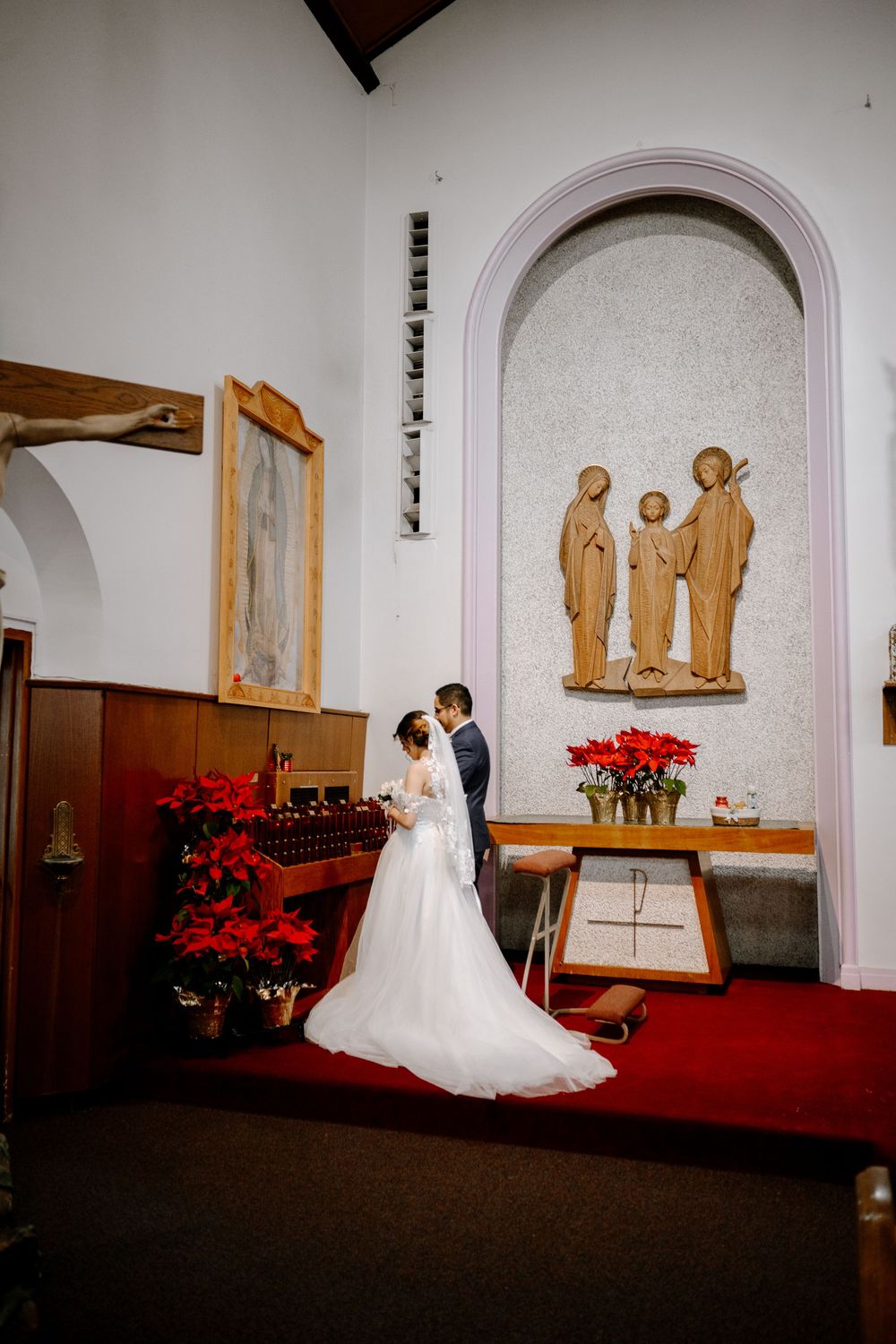 Bride and groom share a romantic moment at a church altar adorned with red poinsettias and a religious sculpture.