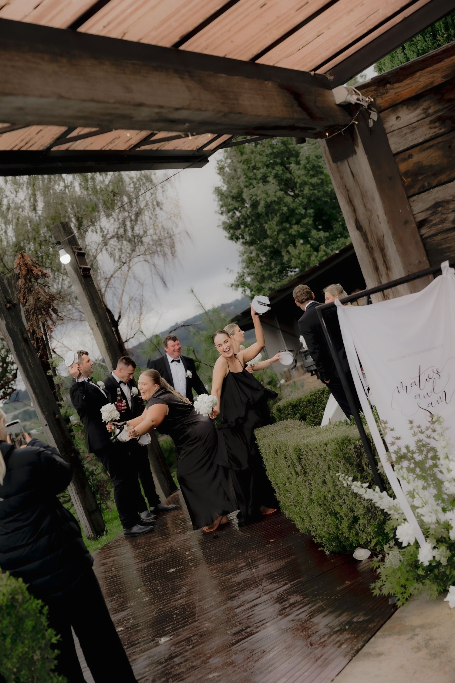 Wedding party in black attire celebrating outdoors under a rustic wooden pergola on a rainy day.