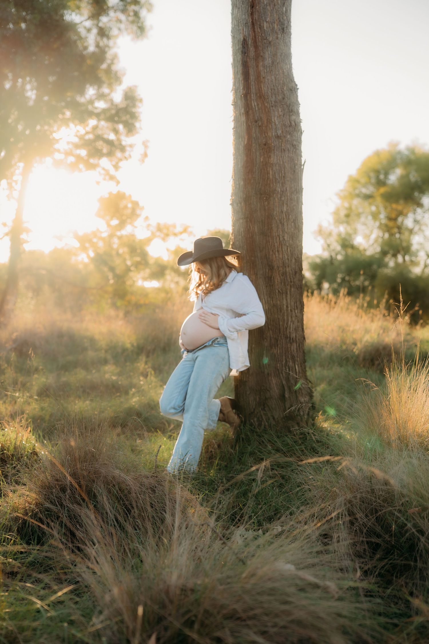 Pregnant woman in cowboy hat and jeans leaning against tree in golden sunset light in a grassy field.