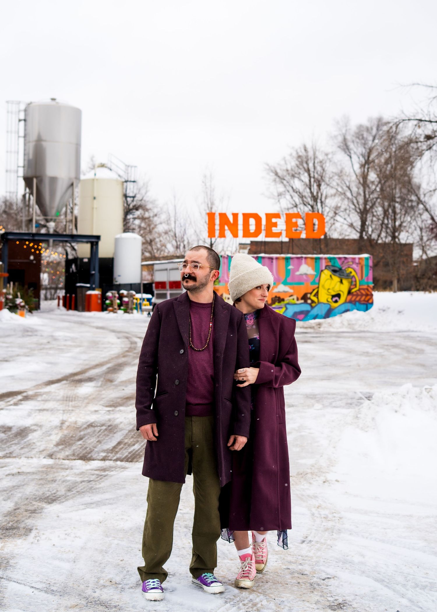 Couple in matching burgundy coats posing in snowy urban setting with colorful INDEED sign backdrop.
