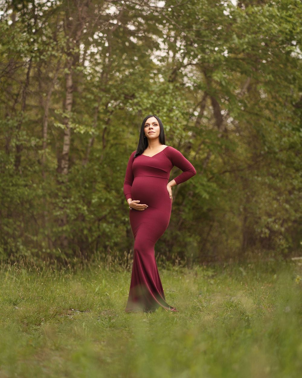 Pregnant woman in burgundy maternity gown standing in a lush green forest clearing during an outdoor maternity photoshoot.