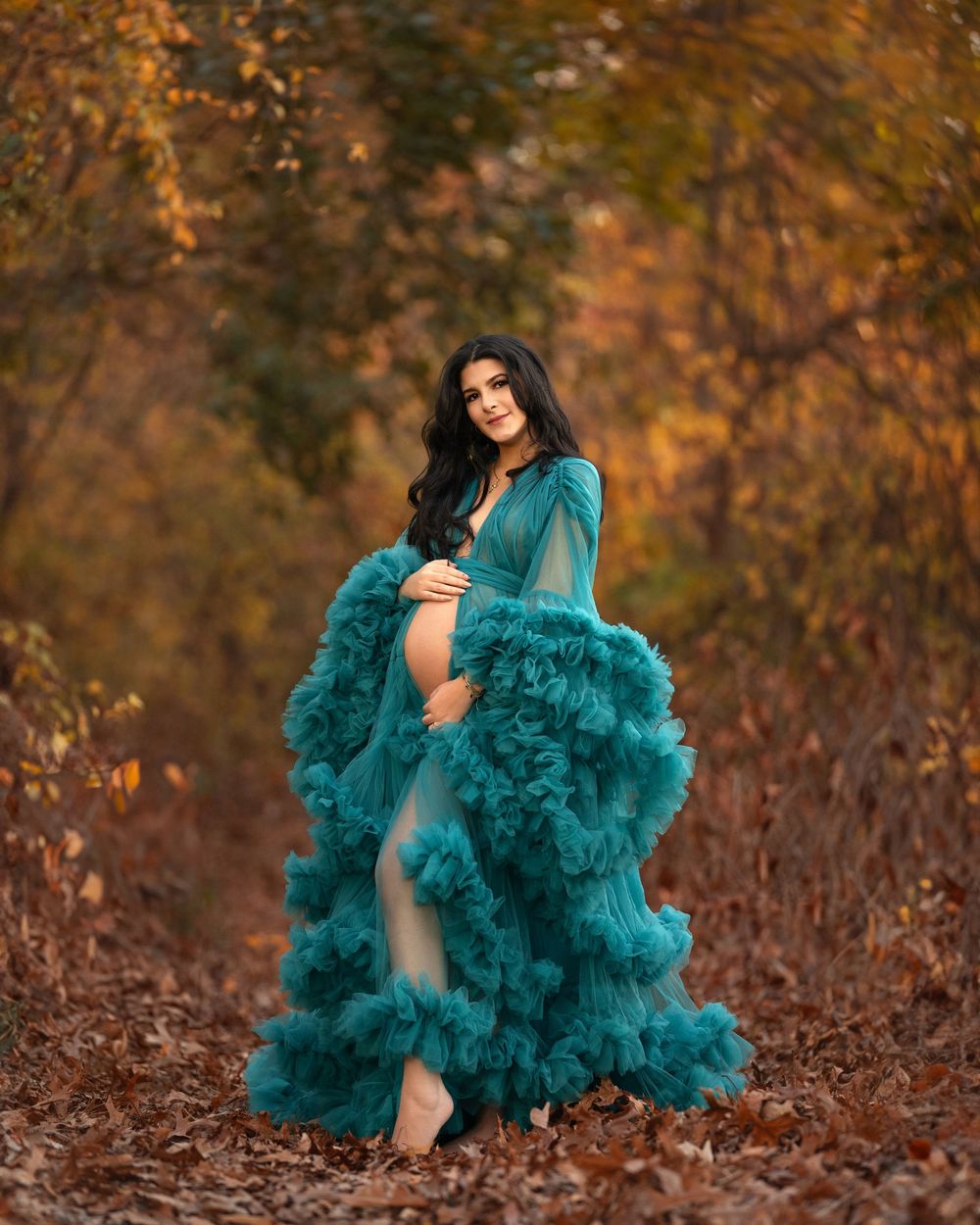Pregnant woman in teal ruffled maternity gown posing outdoors among autumn foliage in a forest path.
