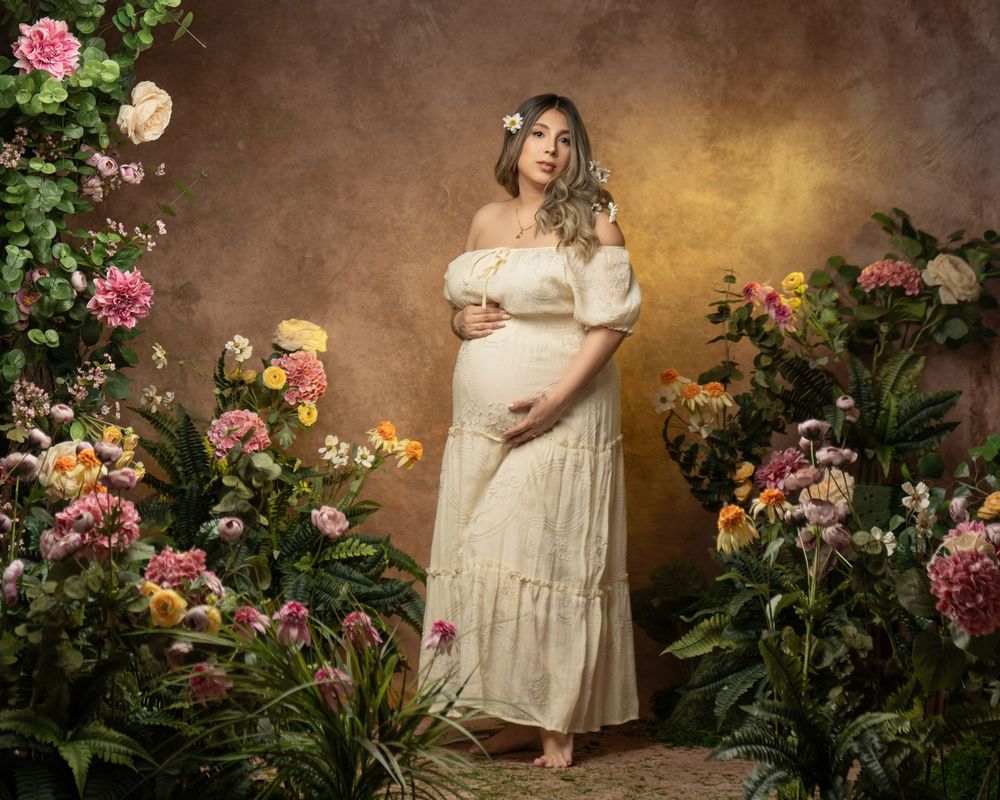 Pregnant woman in white off-shoulder dress poses among colorful flowers in a elegant maternity portrait.