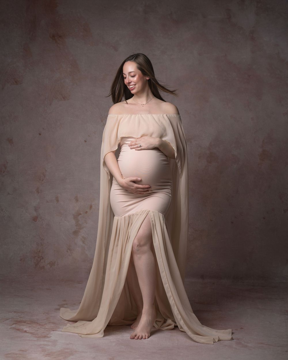 Smiling pregnant woman in elegant beige maternity gown with flowing cape, cradling baby bump in studio portrait.