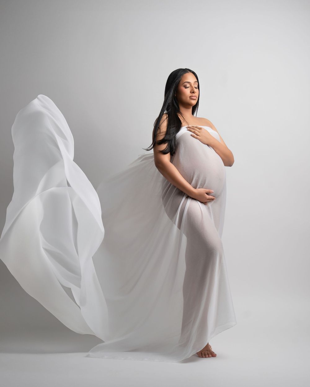 Pregnant woman in flowing white sheer fabric gown poses gracefully in studio maternity photography session.