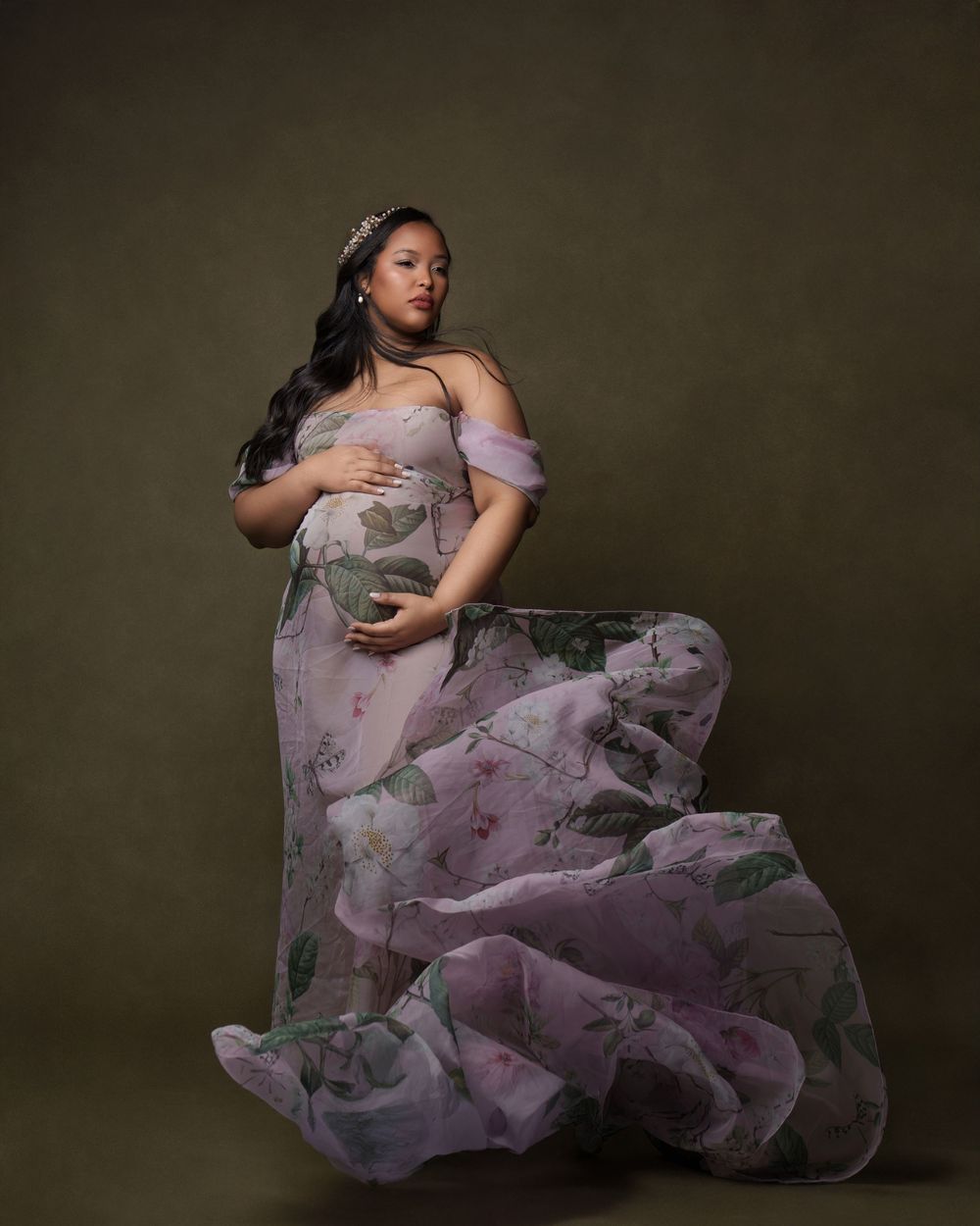 Pregnant woman in floral lavender off-shoulder gown with flowing skirt and tiara poses gracefully in studio.