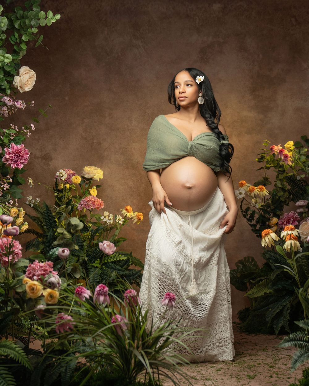 Pregnant woman in green crop top and white lace skirt poses gracefully among colorful flowers in a studio maternity portrait.