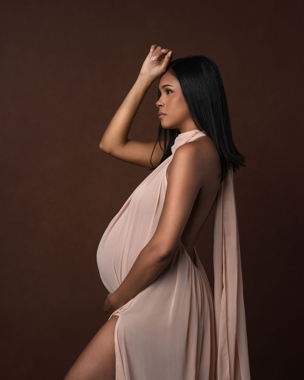 Pregnant woman in elegant backless blush draped gown poses gracefully against a warm brown studio background.