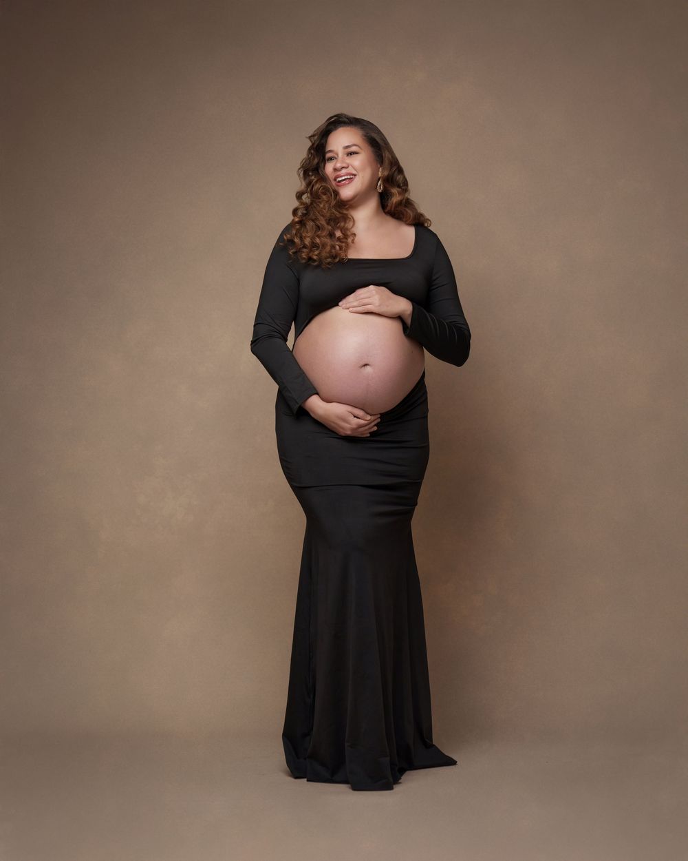 Pregnant woman in black form-fitting maternity gown cradling baby bump, smiling, studio portrait.