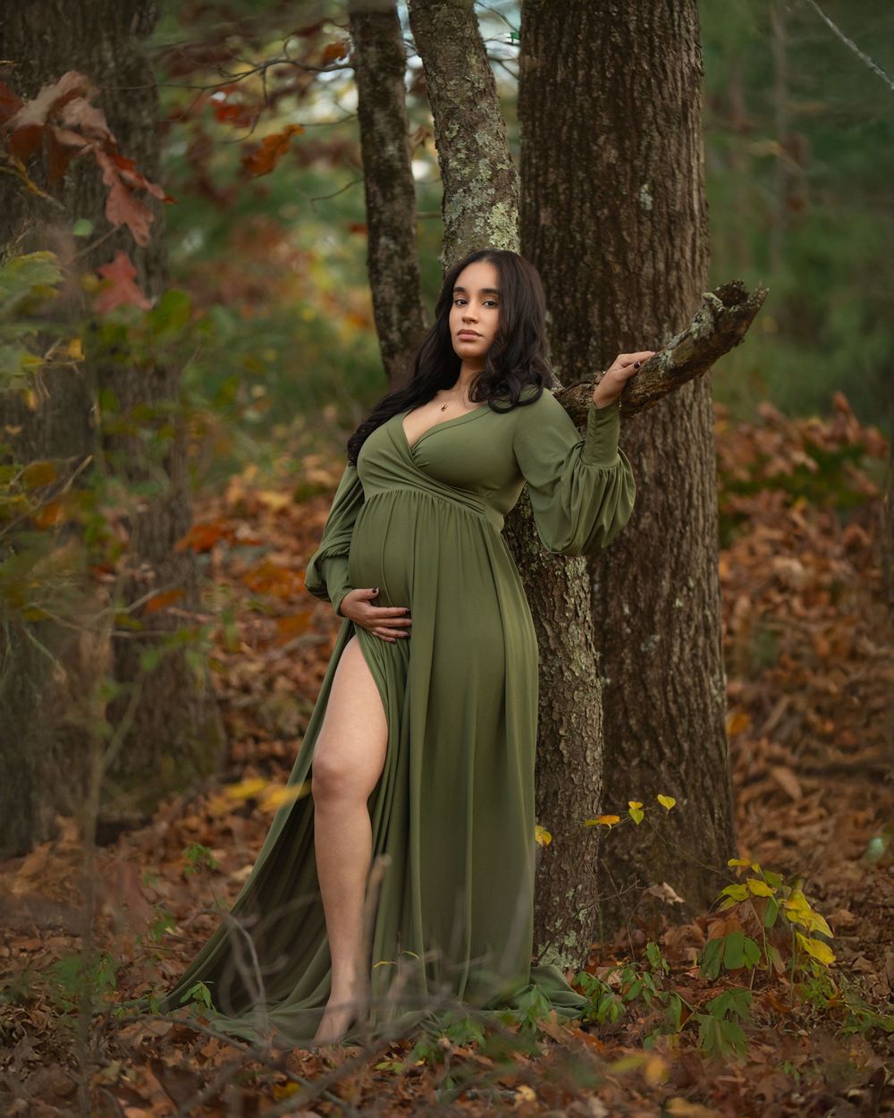 Pregnant woman in olive green maternity gown leaning against a tree in an autumn forest setting.