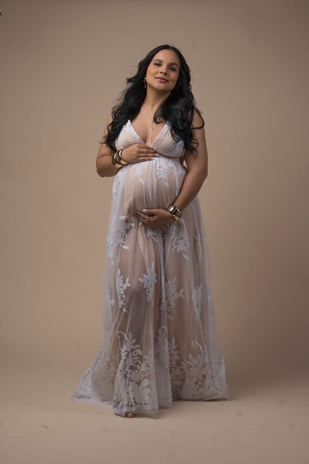 Pregnant woman in sheer lace maternity gown cradling baby bump in studio portrait.