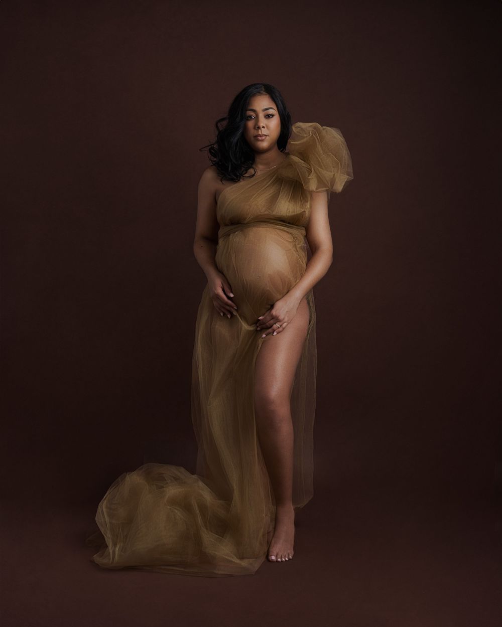 Pregnant woman in elegant gold tulle maternity gown posing gracefully against a warm brown studio backdrop.