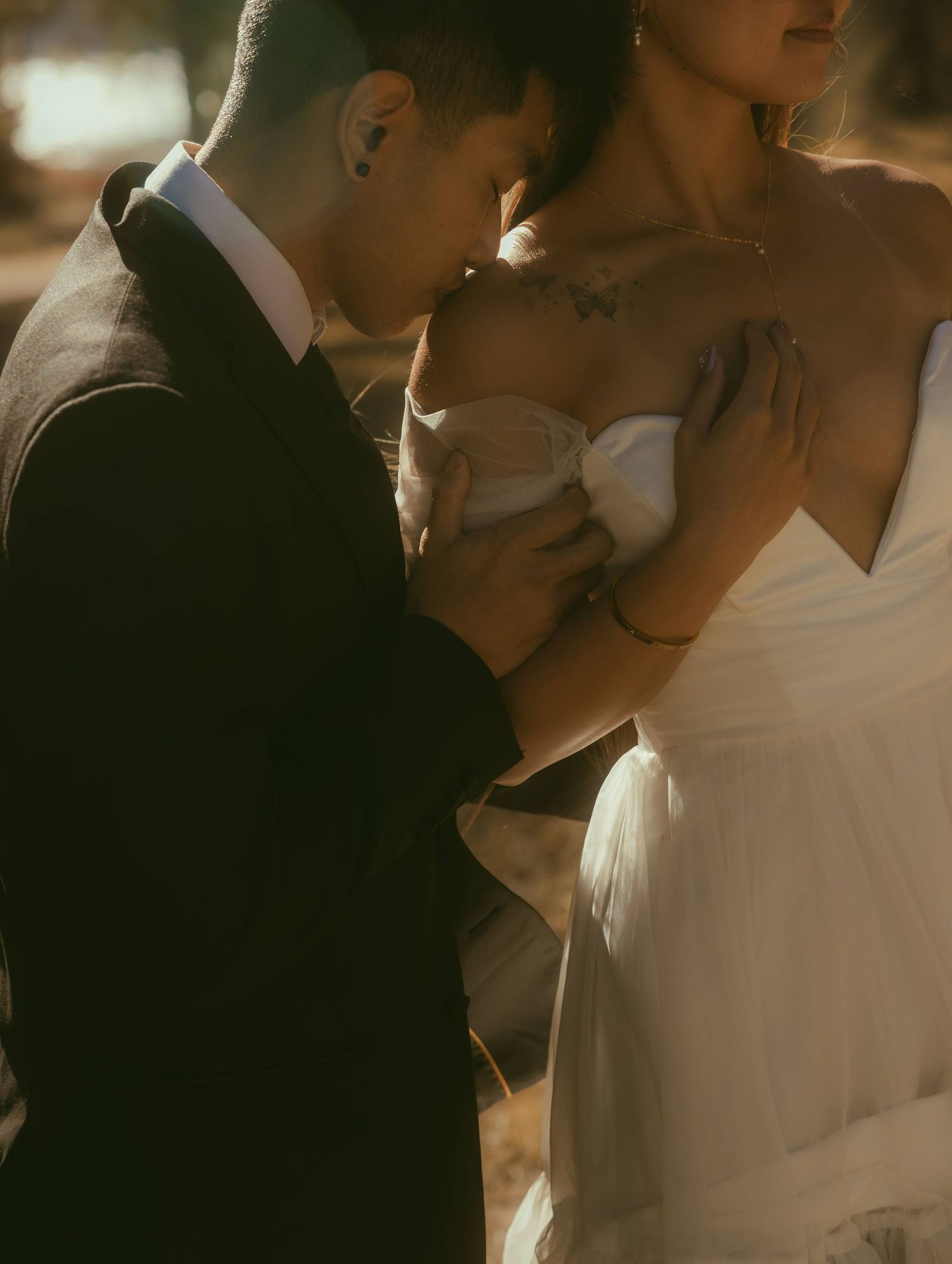 Romantic Los Angeles wedding couple embracing in golden light, groom in dark suit, bride in white strapless gown with floral tattoo.