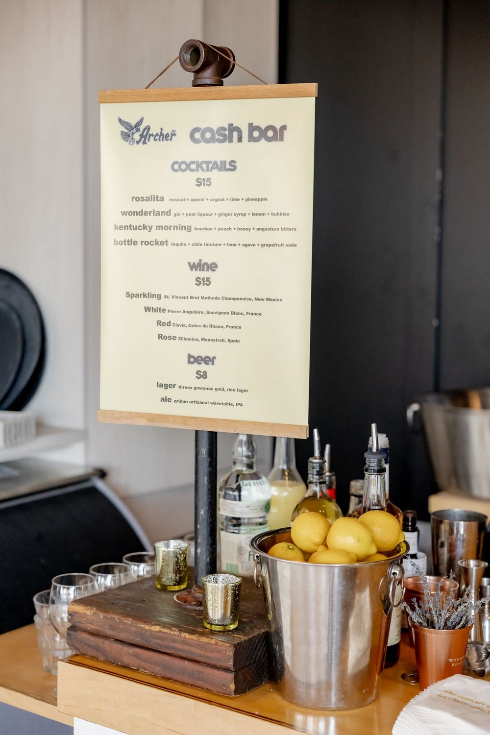 Cash bar menu display with cocktails, wine, and beer pricing, surrounded by lemons, bottles, and bar tools on a wooden counter.