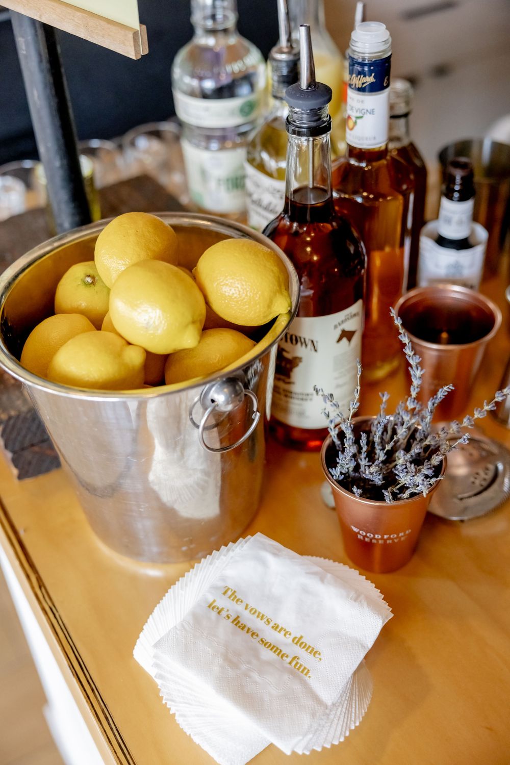 A bar cart with lemons in a silver bucket, liquor bottles, copper mugs, and custom cocktail napkins on a wooden surface.