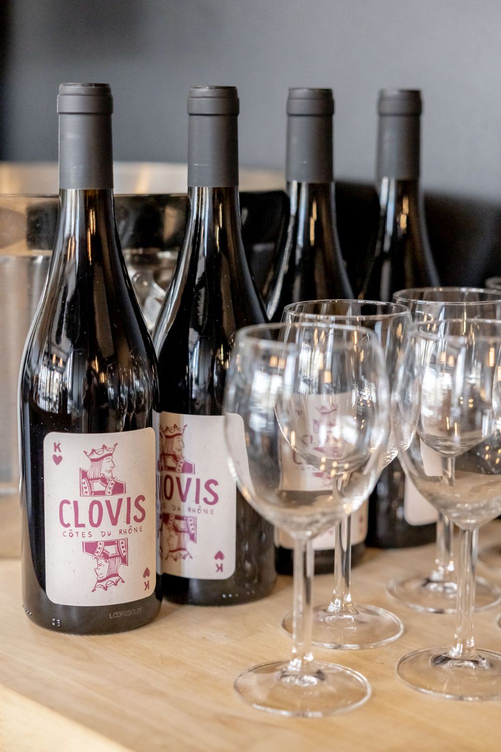 Clovis Cotes du Rhone red wine bottles displayed alongside empty wine glasses on a wooden table.