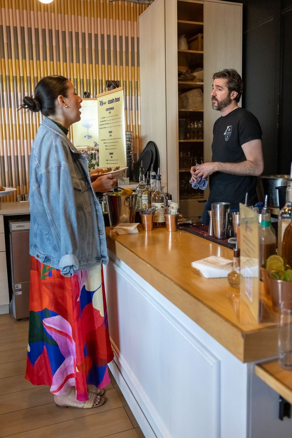 A woman in a colorful skirt and denim jacket orders drinks from a bartender at a modern wooden bar counter.