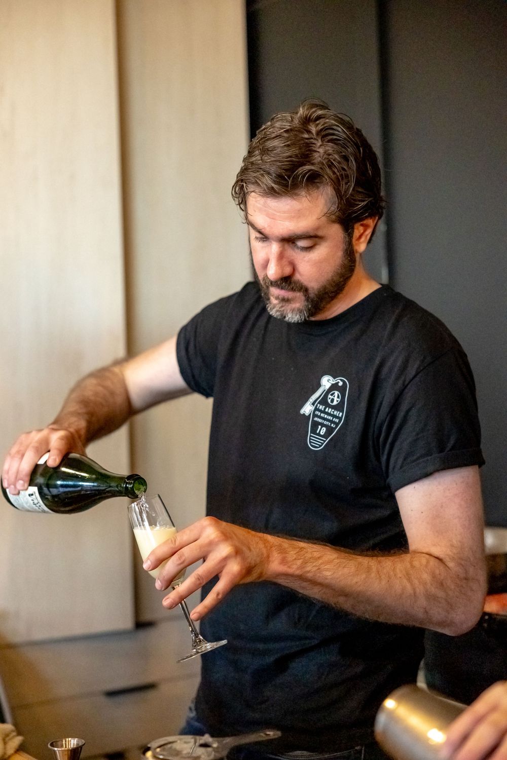 A bearded bartender in a black t-shirt carefully pours sparkling wine into a champagne flute at a modern bar.