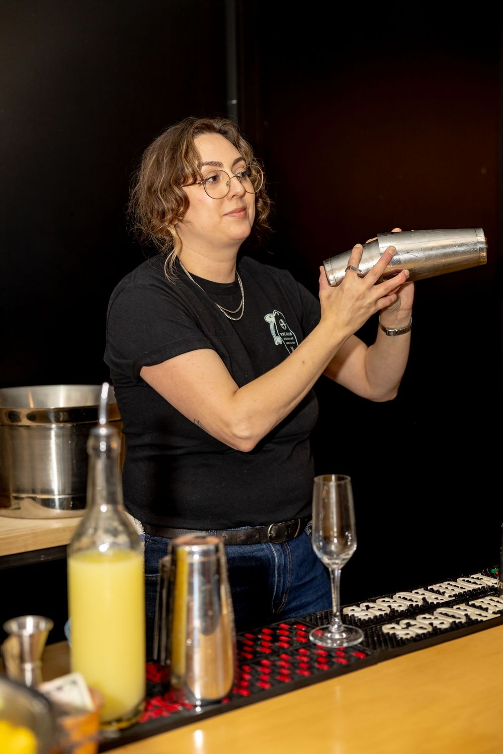 Bartender shaking a cocktail mixer behind a bar with glasses and bottles in foreground.