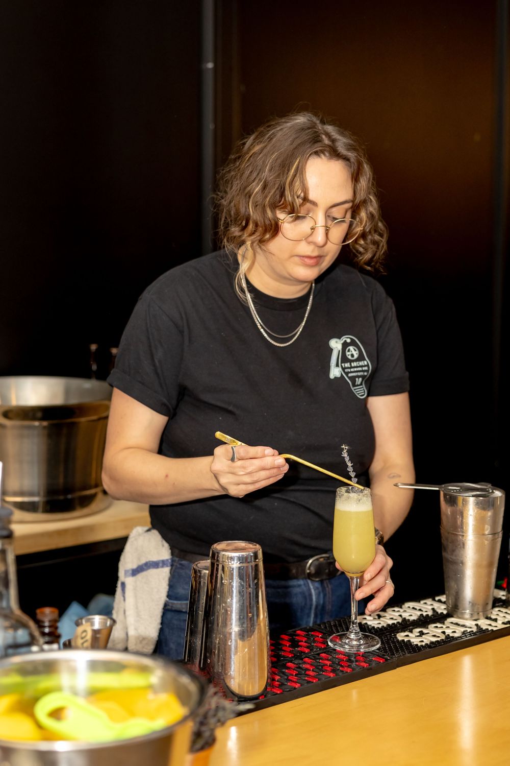 Female bartender in black t-shirt garnishing a frothy cocktail at a bar counter with shakers nearby.