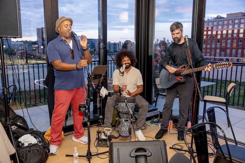 Three musicians perform indoors, one singing, one seated, one playing guitar, with a city skyline visible through large windows.