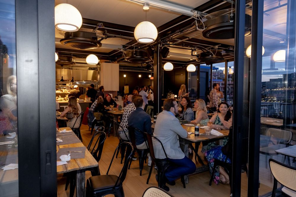 Busy restaurant patio with diners seated at tables under warm pendant lights in a modern indoor-outdoor space.
