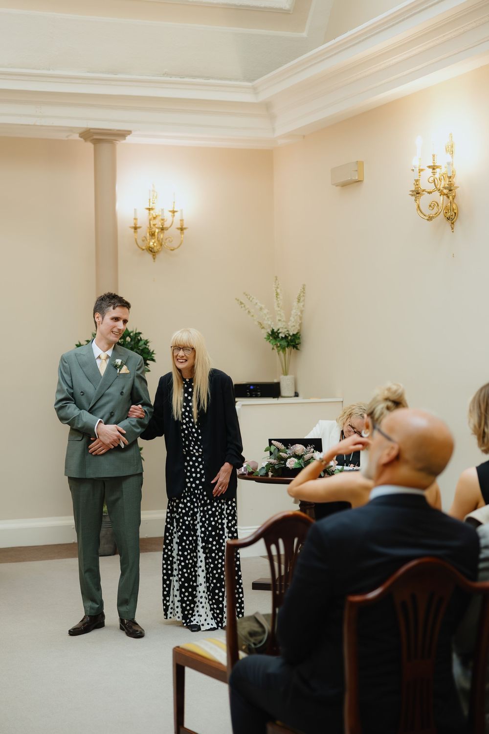 Groom in green suit stands with officiant during intimate wedding ceremony with seated guests.