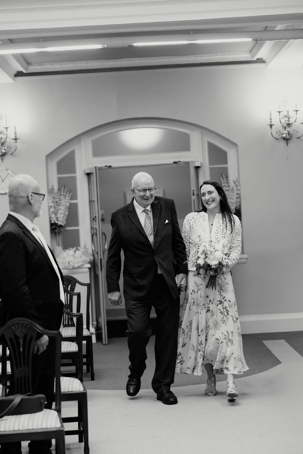 Bride in floral dress walks down aisle escorted by father in black and white wedding photo.