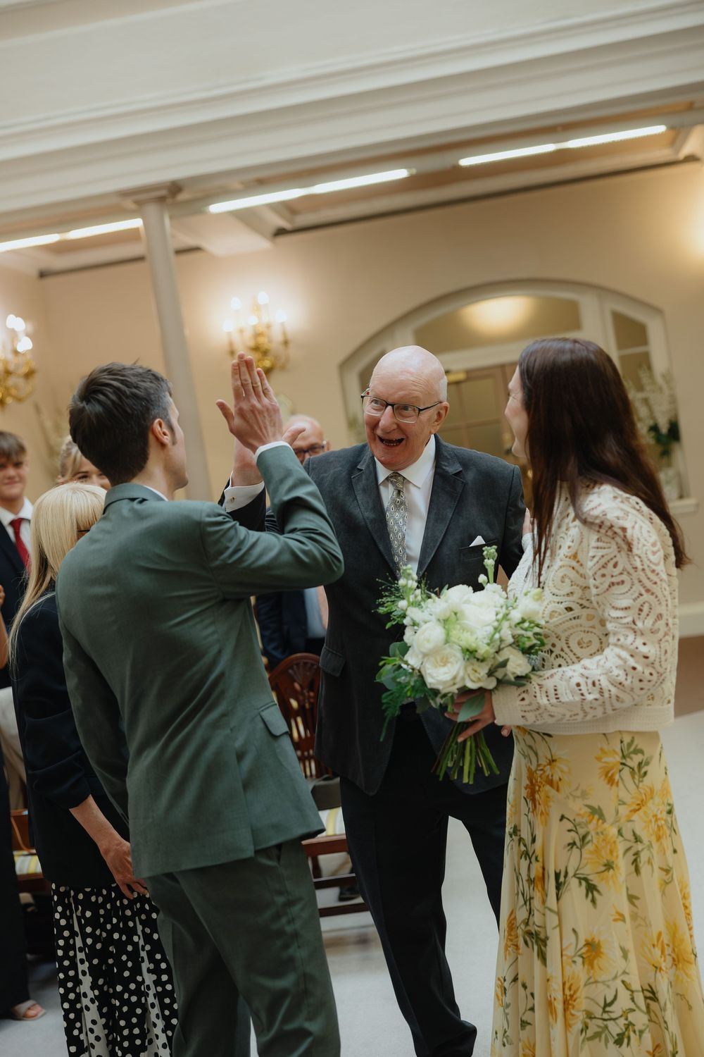 Groom in green suit high-fiving father of bride during wedding ceremony while bride holds white floral bouquet.