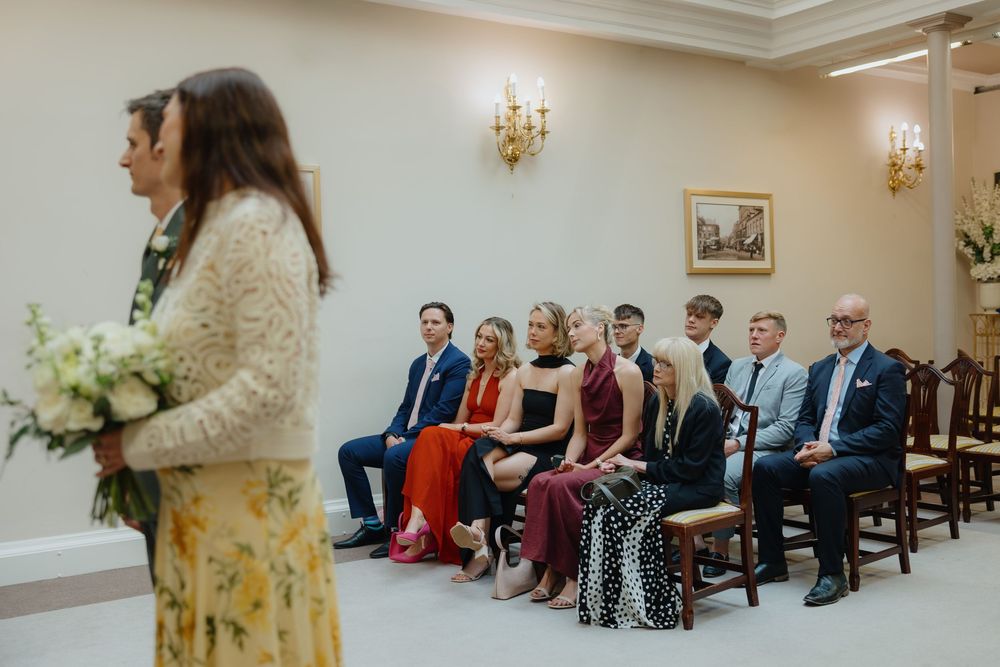 Wedding guests seated in elegant ceremony room watching bride holding white floral bouquet.