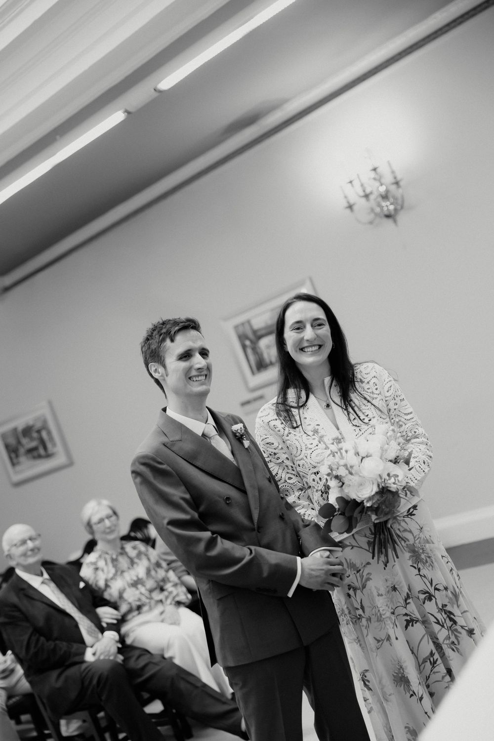 Black and white photo of smiling bride and groom standing together during their wedding ceremony holding bouquet.