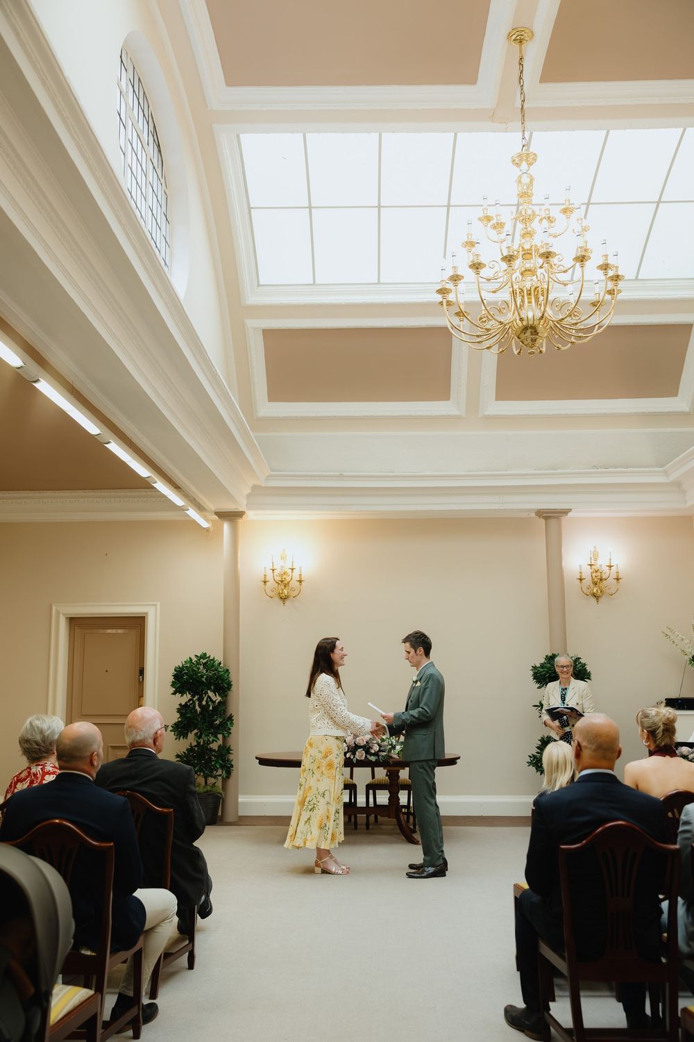 Couple exchanges vows in elegant wedding ceremony room with gold chandelier and seated guests.