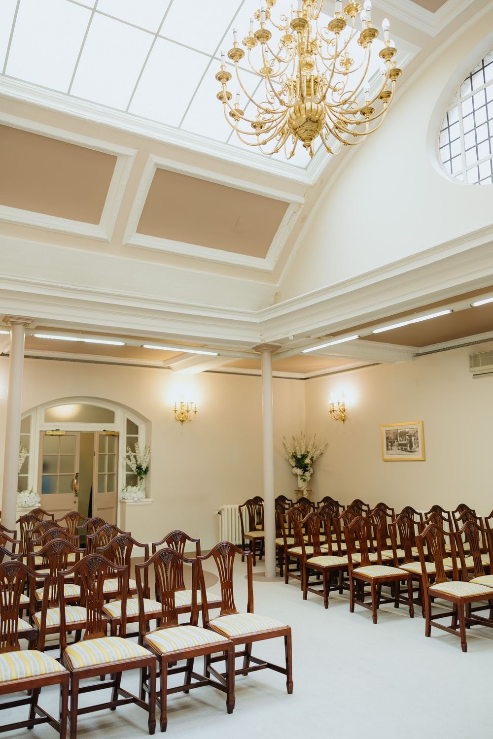 Elegant wedding ceremony room with wooden chairs, gold chandelier, and skylight ceiling in a classic venue.