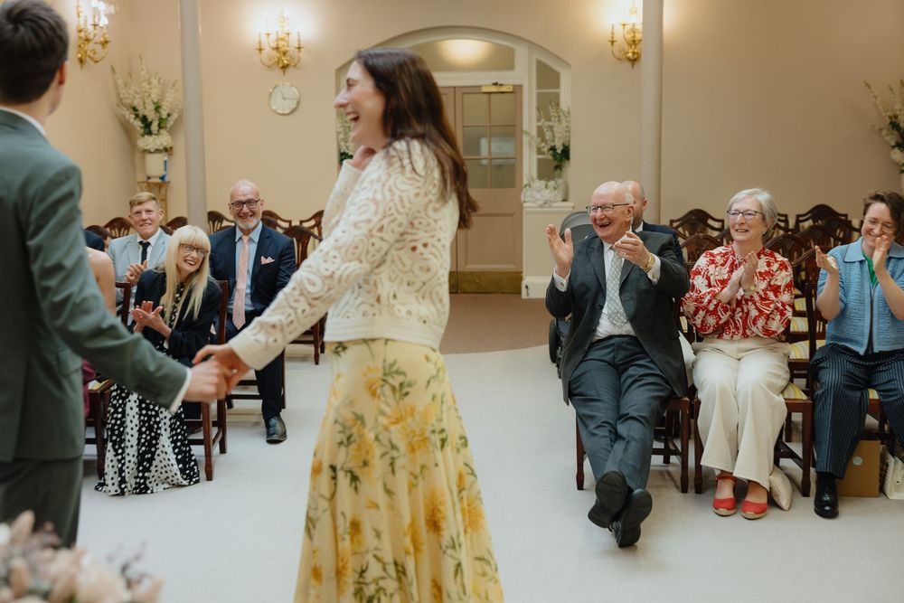 Joyful woman in floral skirt dancing at elegant wedding ceremony while guests clap and celebrate in bright venue.